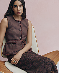 A woman wearing a brown textured vest and lace skirt sits in a modern chair against a plain pink background. carousel Product Image 4
