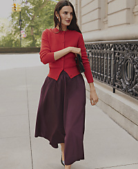 A woman wearing a red cardigan and a long purple skirt walks along a city sidewalk next to an ornate black iron fence. carousel Product Image 4