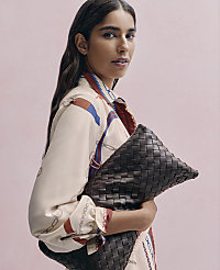 A woman in a patterned cream blouse holds a large woven brown leather clutch against a solid pink background. carousel Product Image 3