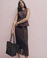 A woman wearing a maroon textured vest and lace skirt holds a large dark tote bag against a plain pink background. carousel Product Image 3