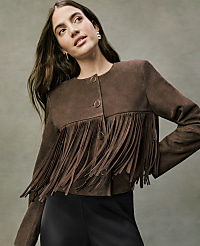 A woman wearing a brown suede fringe jacket and black pants poses against a neutral, textured grey background. carousel Product Image 1