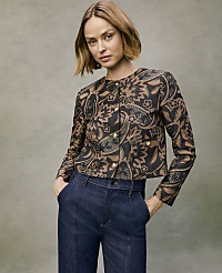 A woman wearing a patterned brown and black jacket and dark blue jeans stands against a neutral, textured grey background. carousel Product Image 1