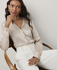 A woman wearing a champagne-colored satin blouse and white trousers sits in a wooden chair against a plain light gray background. carousel Product Image 1