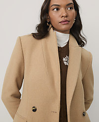A woman wearing a tan coat over a brown sweater and white turtleneck stands against a plain, light-colored background. carousel Product Image 4