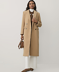 A woman wearing a long camel-colored coat, white trousers, and brown shoes stands against a plain white background. carousel Product Image 1