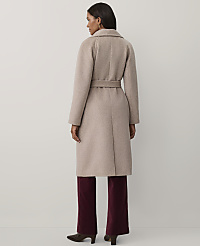 A woman wearing a long beige belted coat and burgundy trousers stands with her back to the camera against a plain light grey background. carousel Product Image 2