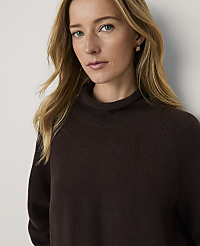 A blonde woman wearing a dark brown turtleneck sweater and blue jeans sits against a plain, light-colored background. carousel Product Image 3