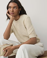 A woman in a shimmering cream sweater and white trousers holds a gold clutch against a plain light gray background. carousel Product Image 1