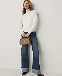 A woman wearing a white turtleneck sweater and wide-leg blue jeans holds a brown handbag against a plain white background. carousel Product Image 3