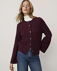 A woman wearing a burgundy cable-knit cardigan over a white turtleneck and blue jeans stands against a plain, light-colored background. carousel Product Image 1