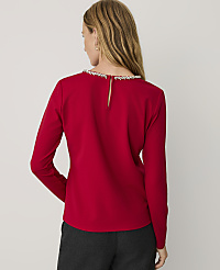 A woman wearing a red long-sleeved top with an embellished neckline and dark grey trousers stands against a plain light grey background. carousel Product Image 2