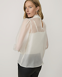 A woman wearing a sheer white high-neck blouse and black leather pants stands against a plain light-colored background. carousel Product Image 2