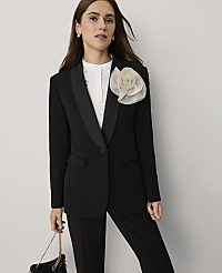 A woman wearing a black suit with a white floral accent and a white shirt stands against a plain light gray background. carousel Product Image 4