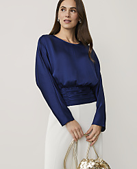 A woman wearing a navy blue satin top and white trousers holds a gold woven bag against a plain light grey background. carousel Product Image 1
