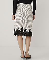 A person stands against a plain white background wearing a black long-sleeved top and a white patterned skirt with black lace trim. carousel Product Image 3
