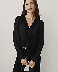 A woman wearing a black lace-detailed blouse and black trousers stands against a plain, light-colored background. carousel Product Image 1