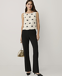 A woman wearing a patterned cream top and black trousers stands against a plain white background, holding a gold woven handbag. carousel Product Image 3