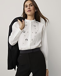 A woman wearing a white embellished shirt and black trousers stands against a plain light grey background, holding a black blazer over her shoulder. carousel Product Image 1
