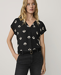 A blonde woman wearing a black floral print blouse and black jeans stands against a plain, light-colored background. carousel Product Image 1