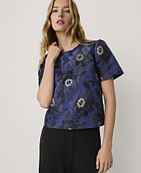 A woman wearing a blue floral patterned top and black trousers stands against a plain light grey background. carousel Product Image 1