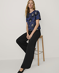 A woman wearing a blue floral top and black trousers sits on a wooden stool against a plain white background. carousel Product Image 3