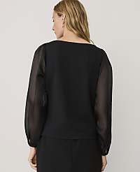 A woman wearing a black top with sheer sleeves stands with her back to the camera against a plain light gray background. carousel Product Image 2