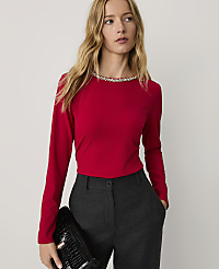 A woman wearing a red long-sleeved top with an embellished neckline and grey trousers stands against a plain white background. carousel Product Image 1