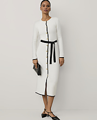 A woman wearing a white long-sleeved dress with black trim and a black belt stands against a plain white background. carousel Product Image 1