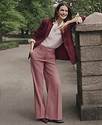 A smiling woman in a burgundy blazer and pink wide-leg trousers leans against a stone pillar on a paved path surrounded by lush green trees. carousel Product Image 4