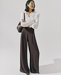 A woman wearing a white lace-detailed blouse and wide-leg brown trousers stands with her arms crossed against a plain, light-colored background. carousel Product Image 5