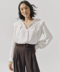 A woman wearing a white lace-collared blouse and brown trousers stands against a plain light gray background. carousel Product Image 4