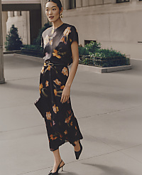 A woman wearing a floral black dress and heels walks along a city sidewalk with greenery and a building in the background. carousel Product Image 3