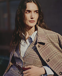A woman wearing a plaid blazer and light blue shirt poses against a blurred, dark architectural background. carousel Product Image 7