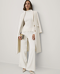 A woman wearing an all-white ensemble consisting of a ribbed sweater, wide-leg trousers, and a long coat stands against a plain white wall. carousel Product Image 1