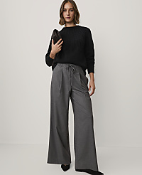 A woman wearing a black cable-knit sweater and wide-leg grey trousers stands against a plain white background. carousel Product Image 1
