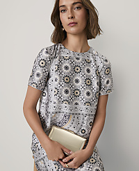 A woman wearing a grey patterned dress and holding a gold clutch stands against a plain white background. carousel Product Image 3