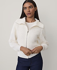 A woman wearing a white textured zip-up sweater and dark blue jeans stands against a plain, light-colored background. carousel Product Image 1