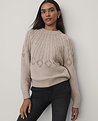 A woman wearing a beige cable-knit sweater and black jeans stands against a plain, light-colored wall. carousel Product Image 1