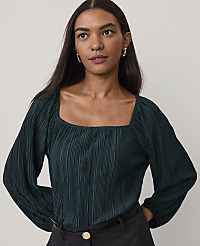 A woman wearing a dark green pleated blouse and grey trousers stands against a plain, light-colored background. carousel Product Image 1