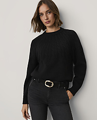 A woman wearing a black cable-knit sweater and black jeans with a gold buckle belt stands against a plain white wall. carousel Product Image 1