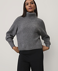 A woman wearing a grey ribbed turtleneck sweater and black trousers stands against a plain light grey background. carousel Product Image 1