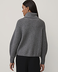 A woman wearing a grey ribbed turtleneck sweater and black trousers stands against a plain light-colored background. carousel Product Image 2