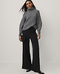 A woman wearing a grey ribbed turtleneck sweater and wide-leg black trousers stands against a plain white background. carousel Product Image 3
