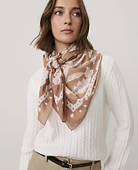 A woman wearing a white cable-knit sweater and a brown patterned scarf stands against a plain white background. carousel Product Image 1