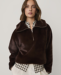 A woman wearing a dark brown faux-fur jacket over a checkered shirt and white trousers stands against a plain light gray background. carousel Product Image 1