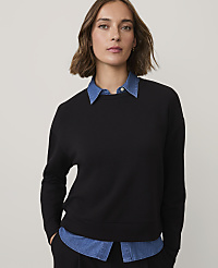 A woman wearing a black sweatshirt over a blue collared shirt stands against a plain, light-colored background. carousel Product Image 1