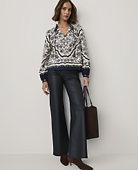 A woman wearing a patterned blouse and navy leather trousers stands against a plain white background while holding a brown tote bag. carousel Product Image 3