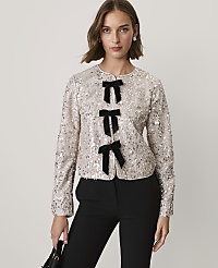 A woman wearing a sequined champagne-colored jacket with black bow accents and black trousers stands against a plain light gray background. carousel Product Image 1