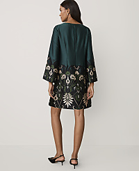 A woman stands with her back to the camera wearing a dark green dress with an ornate floral pattern at the hem against a plain, light-colored background. carousel Product Image 2