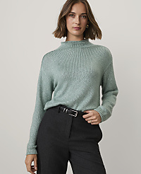 A woman wearing a sage green knit sweater and dark trousers stands against a plain, light-colored background. carousel Product Image 1